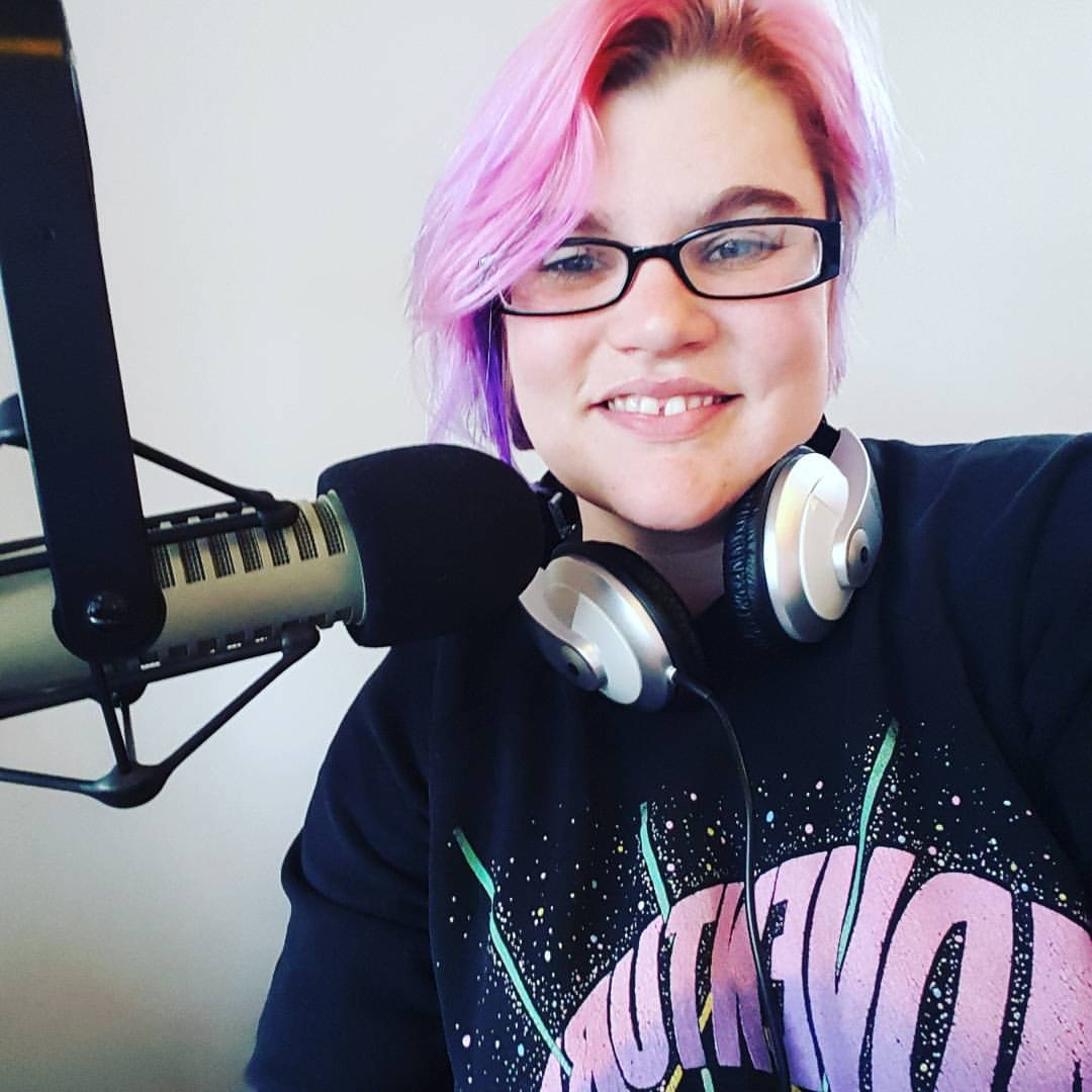 Woman with glasses wears headphones around neck and sits behind a broadcast microphone